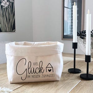 K&ouml;nnte beinhalten: Ein wei&szlig;er Stoffkorb mit schwarzem Text, der "Viel Gl&uuml;ck im neuen Zuhause!" lautet. Der Korb steht auf einem Holztisch mit zwei schwarzen Kerzenst&auml;ndern und wei&szlig;en Kerzen.