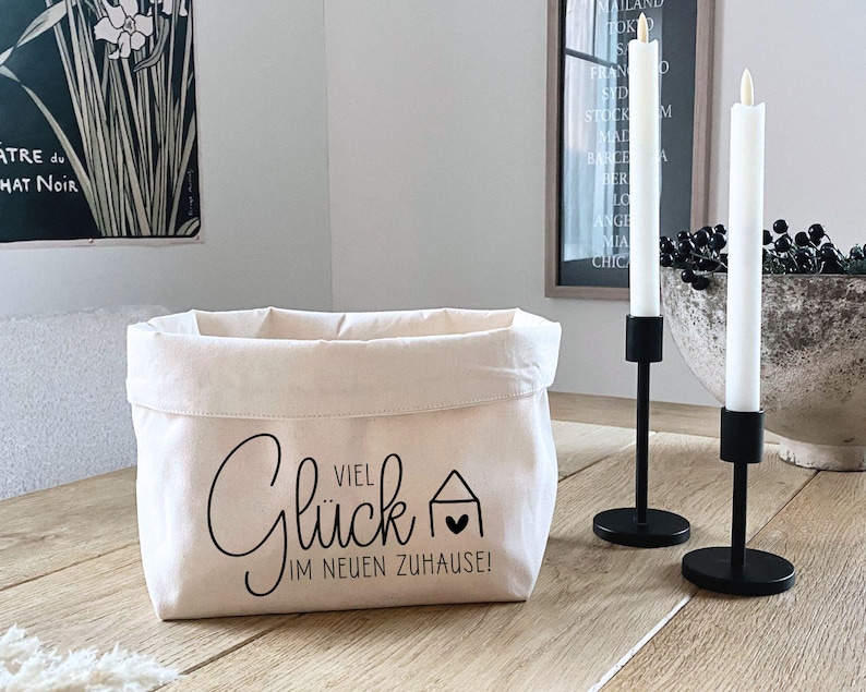 K&ouml;nnte beinhalten: Ein wei&szlig;er Stoffkorb mit schwarzem Text, der "Viel Gl&uuml;ck im neuen Zuhause!" lautet. Der Korb steht auf einem Holztisch mit zwei schwarzen Kerzenst&auml;ndern und wei&szlig;en Kerzen.