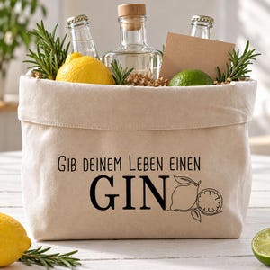 Könnte beinhalten: Ein beigefarbener Stoffkorb mit dem schwarzen Aufdruck "GIB DEINEM LEBEN EINEN GIN" und einer Strichzeichnung einer Zitrone. Der Korb enthält klare Glasflaschen, eine gelbe Zitrone, eine Limette und Zweige von grünem Rosmarin.