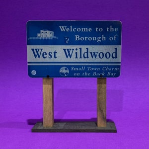 May include: A blue and white sign with the text "Welcome to the Borough of West Wildwood" and "Small Town Charm on the Back Bay". The sign is mounted on two wooden posts.