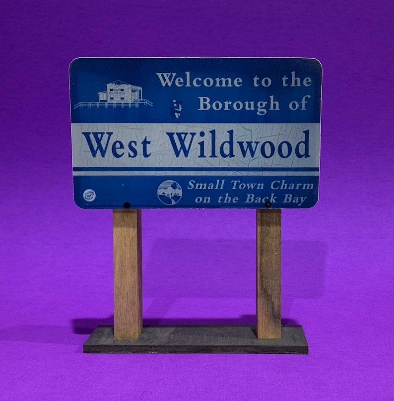 May include: A blue and white sign with the text "Welcome to the Borough of West Wildwood" and "Small Town Charm on the Back Bay". The sign is mounted on two wooden posts.