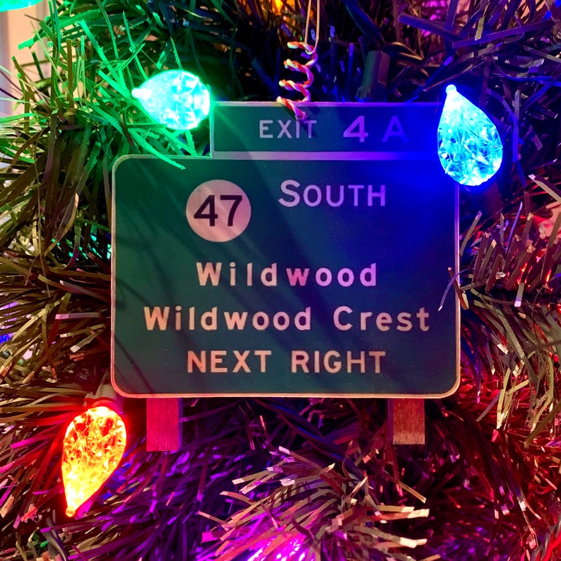 May include: A green and white highway sign with the text "Exit 4A South" and "47" in a circle. The sign also reads "Wildwood Wildwood Crest Next Right". The sign is hanging on a Christmas tree with colorful lights.