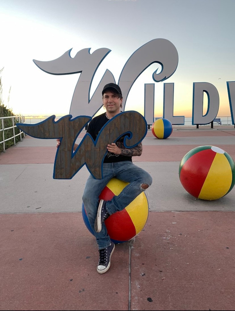 Pode incluir: Uma pessoa sentada em uma grande bola de praia, segurando uma placa de madeira com a letra "W" em azul e branco. A placa est&aacute; na frente de uma grande placa branca que diz "WILDWOOD".