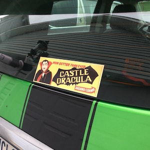May include: A green car with black racing stripes and a sticker that says "Now Better Than Ever Castle Dracula Around The Corner".