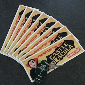 May include: A stack of yellow and red signs with the text "Now Better Than Ever" and "Castle Dracula" with a black and white illustration of a vampire. The signs are on a dark gray surface with a green key chain with a motel key and the text "Bates Motel" on it.