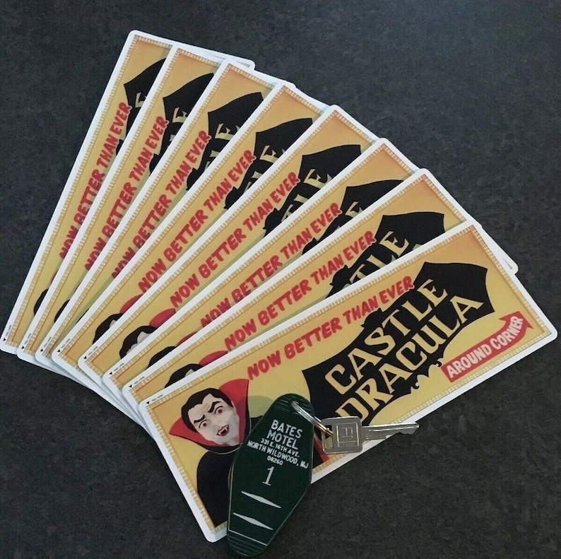 May include: A stack of yellow and red signs with the text "Now Better Than Ever" and "Castle Dracula" with a black and white illustration of a vampire. The signs are on a dark gray surface with a green key chain with a motel key and the text "Bates Motel" on it.