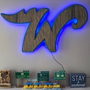 Pode incluir: Uma placa de madeira com um contorno de n&eacute;on azul em forma da letra "W". Abaixo da placa, h&aacute; v&aacute;rios sinais de estrada em miniatura com o texto "North Wildwood", "Wildwood" e "N Cape May". Um pequeno sinal de madeira com o texto "STAY anchored" est&aacute; &agrave; direita.