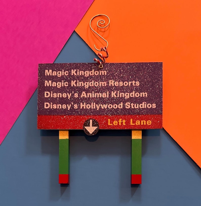 May include: A purple and red sign with glitter that reads "Magic Kingdom", "Magic Kingdom Resorts", "Disney's Animal Kingdom", and "Disney's Hollywood Studios". The sign also has a white arrow pointing down and the words "Left Lane" in white text on a red background.