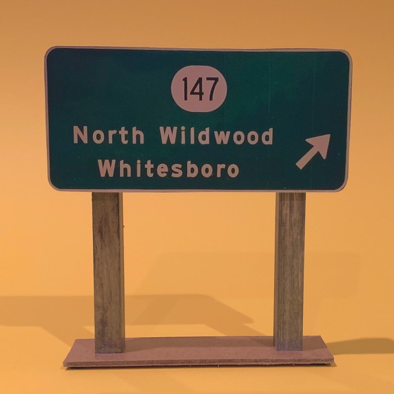 May include: A green road sign with white lettering and a white circle with the number 147 inside. The sign reads "North Wildwood Whitesboro" with a white arrow pointing to the right.