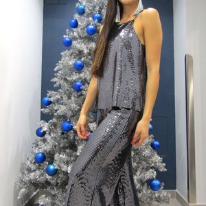 Pode incluir: Uma mulher vestindo um top e calças de lantejoulas prateadas está em frente a uma árvore de Natal prateada com enfeites azuis. Ela está usando brincos prateados e sandálias pretas.