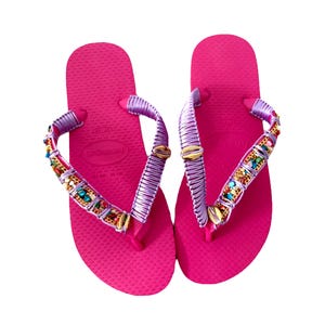 Op de afbeelding: Een paar roze slippers met paarse bandjes en kleurrijke kralenversieringen. De bandjes zijn versierd met gouden accenten en kleine schelpen.