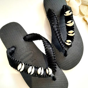 Op de afbeelding: Zwarte slippers met een zwarte gevlochten band versierd met witte schelpen. De slippers hebben de merknaam "Havaianas" op de band gedrukt.