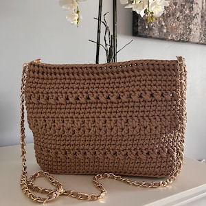 May include: A brown crocheted crossbody bag with a gold chain strap. The bag has a woven texture and a rectangular shape.