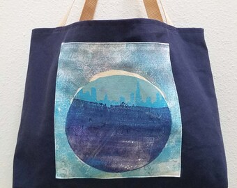 Puede incluir: Una bolsa de lona azul marino con correas de color canela. La bolsa presenta un parche cuadrado con un diseño de ciudad azul y blanco dentro de un círculo.
