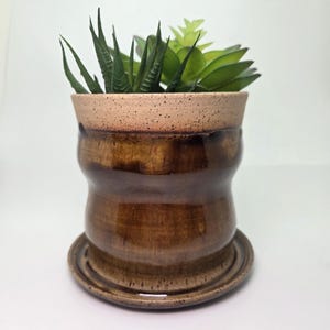 May include: A brown ceramic planter with a matching saucer, holding green and light green succulents. The planter has a unique hourglass shape with a speckled tan rim. The glossy finish gives it a polished look, perfect for indoor plants.