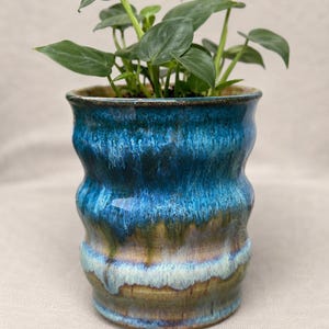 May include: A cylindrical ceramic planter with a wavy, rippled exterior surface. The glaze features a vibrant gradient of deep cobalt blue at the top, transitioning into earthy olive green and creamy white tones near the base. A green plant sits inside.