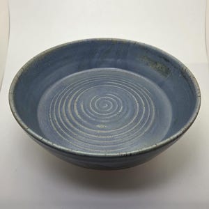 Może przedstawiać: Ręcznie robiona, niebieska ceramiczna miska ze spiralnym wzorem pośrodku. Misa ma szeroki, płytki kształt i teksturowaną powierzchnię. Obręcz miski jest nieco ciemniejsza niż wnętrze. Unikalny element do serwowania lub ekspozycji.