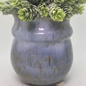 May include: A ceramic pot with a blue and brown glaze, holding a cluster of green, cone-shaped faux plants. The pot has a rounded body, a flared rim, and three small, white feet. The overall aesthetic is rustic and decorative.