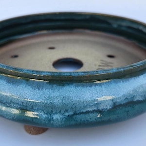 May include: A round ceramic bonsai pot with a glossy teal and blue glaze. The interior is a smooth, light beige color with a central drainage hole and smaller holes around the base. The pot features a raised rim and small, sturdy feet.