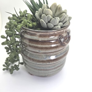 Puede incluir: Una maceta de cerámica con un esmalte marrón y azul, llena de una variedad de suculentas artificiales. La maceta tiene un diseño texturizado y acanalado y pequeñas asas. El arreglo incluye plantas verdes colgantes.