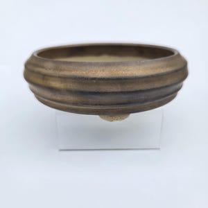 May include: A round, shallow ceramic bonsai pot with a dark, metallic bronze-toned exterior finish. The vessel features a series of horizontal, raised ridges encircling the outer wall and rests on small, light-colored, protruding feet.
