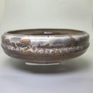 May include: A handcrafted ceramic bowl with a wide, shallow design. The bowl features a textured, earthy aesthetic with a mix of brown, white, and blue-gray glazes. The rim has a raised, decorative band, and the base has a small, rectangular foot.