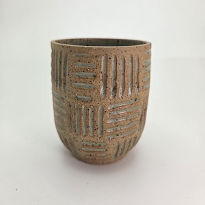 May include: A handmade, textured ceramic cup with a woven pattern. The cup is a warm, earthy brown color with subtle gray accents in the carved design. The cup has a rounded base and a slightly flared rim, perfect for holding beverages.