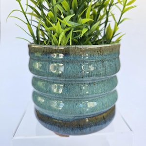 May include: A cylindrical ceramic planter with a ribbed texture and a glossy glaze in shades of teal, blue, and brown. The pot features a dark brown rim and base, with a lighter blue-green center. A small green artificial plant is inside the pot.