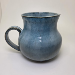 May include: A ceramic mug with a rounded, bulbous body and a curved handle. The mug features a variegated glaze in shades of dusty blue and slate gray, with darker tones concentrated around the rim and handle, creating a smooth, glossy finish.