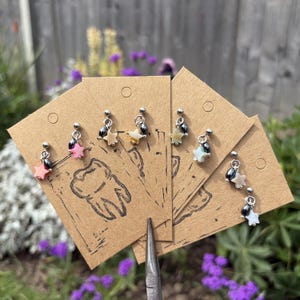 Op de afbeelding: Een collectie zilverkleurige bungelende oorbellen met kleine stervormige bedels in diverse pastelkleuren, gecombineerd met donkere metalen kralen. Elk paar is bevestigd op individuele bruine kraftpapieren kaartjes met een zwarte tand-opdruk.