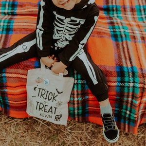 Può includere: Un bambino in costume da scheletro nero, con in mano una borsa di tela con la scritta "Trick or Treat" e il nome "Kevin". Il bambino è seduto su una coperta a quadri.