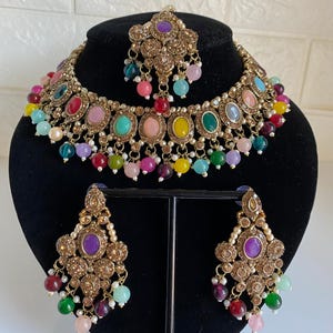 Puede incluir: Un conjunto de joyas dorado con un collar, pendientes y una diadema. El collar y los pendientes tienen detalles de piedras preciosas de colores y cuentas colgantes de varios colores. El conjunto se exhibe sobre una cabeza de maniquí negra.