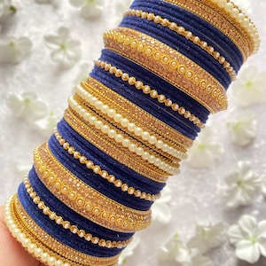 May include: A stack of blue velvet bangles with gold and pearl embellishments. The bangles are stacked on top of each other and are decorated with gold beads, pearls, and glitter.