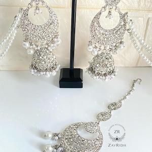 May include: Silver-toned bridal jewelry set featuring a pair of chandelier earrings and a headpiece. The earrings have a crescent design with dangling faux pearls and a bell-shaped bottom. The headpiece has a circular design with faux pearls and the text "ZAYRIDH".