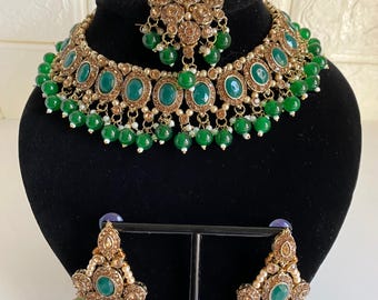May include: A gold-toned jewellery set comprising a necklace, earrings, and a headpiece. The set features oval emerald green stones and small green beads. The necklace and earrings are displayed on a black mannequin head.