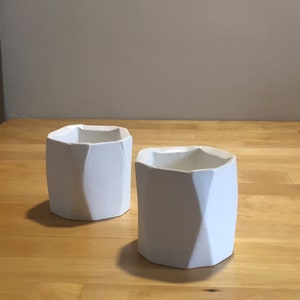 Może przedstawiać: Dwa białe geometryczne doniczki ceramiczne z fakturowaną powierzchnią. Doniczki znajdują się na jasnym drewnianym podłożu.