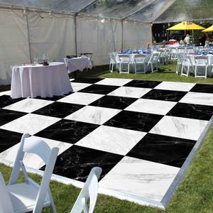 May include: A black and white checkered dance floor set up on a grassy area, surrounded by white chairs and tables with white tablecloths. The scene is set under a tent, with additional tables and umbrellas visible in the background.
