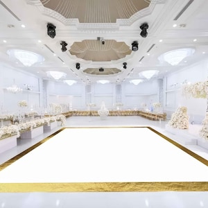May include: A large, white dance floor with a gold border in a white ballroom with white floral arrangements and chandeliers.