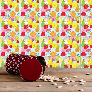 May include: A red and white patterned metal container with a red lid, sitting on a wooden surface with scattered white seeds. The container is in front of a tiled wall with a repeating pattern of fruit and vegetables, including watermelon, strawberries, pears, pineapples, oranges, and cherries.