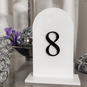 May include: White table number card with a black number 8 on it. The card is shaped like an arch and has a white base.