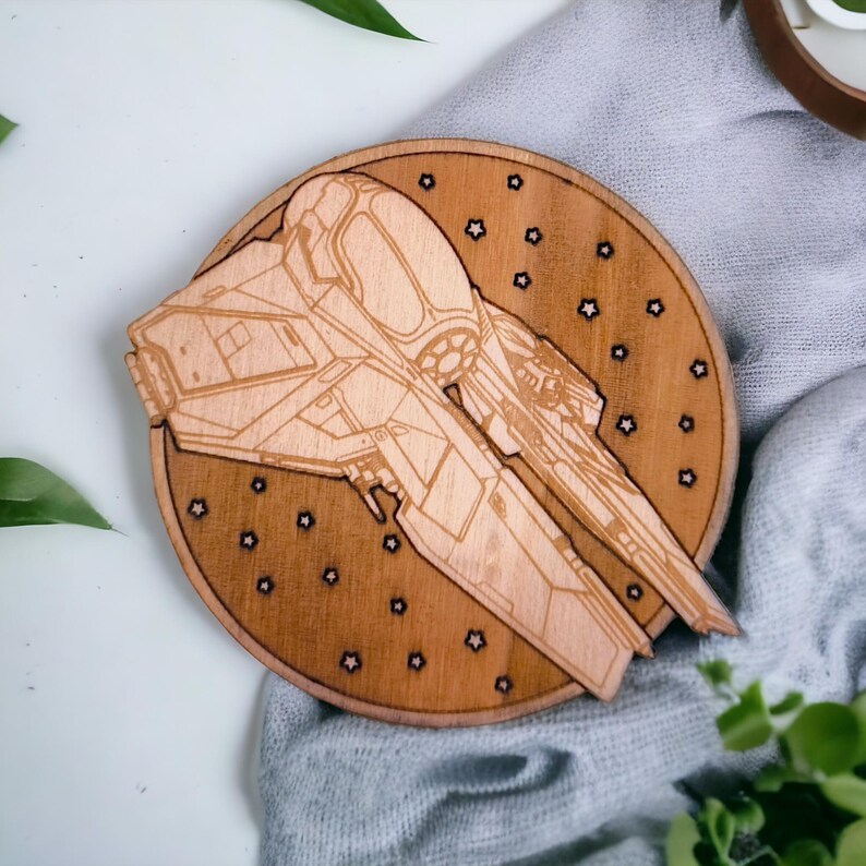 May include: A wooden coaster featuring a detailed laser-cut design of a spaceship. The coaster is round with a dark brown finish and small star-shaped cutouts. The spaceship design is light brown and prominently displayed.