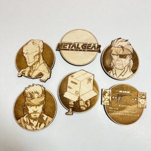 Pode incluir: Uma coleção de seis bases para copos de madeira cortadas a laser com o tema Metal Gear Solid. As bases para copos apresentam retratos de personagens e o logotipo do jogo. Uma base para copos tem o texto "TACTICAL ESPIONAGE ACTION METAL GEAR SOLID".