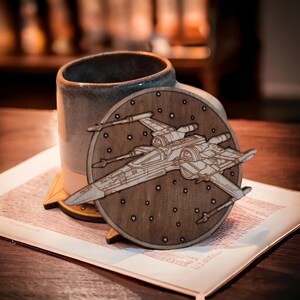 May include: A wooden coaster featuring a detailed laser-cut design of an X-wing starfighter, set against a backdrop of stars. The coaster is round and rests on a wooden stand, next to a ceramic mug.