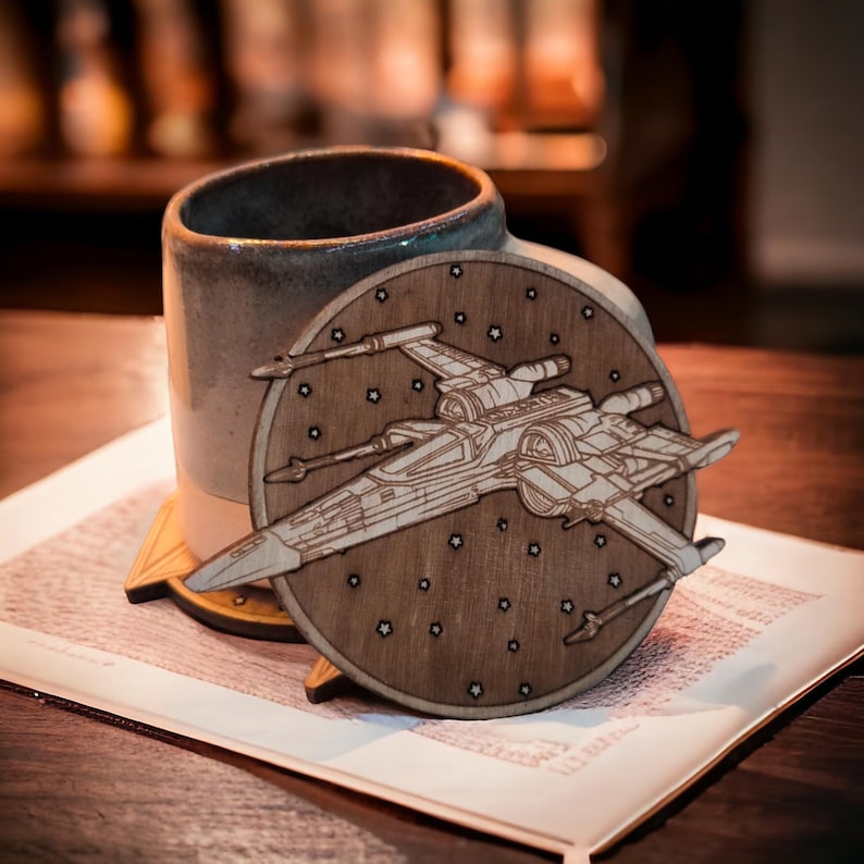 May include: A wooden coaster featuring a detailed laser-cut design of an X-wing starfighter, set against a backdrop of stars. The coaster is round and rests on a wooden stand, next to a ceramic mug.