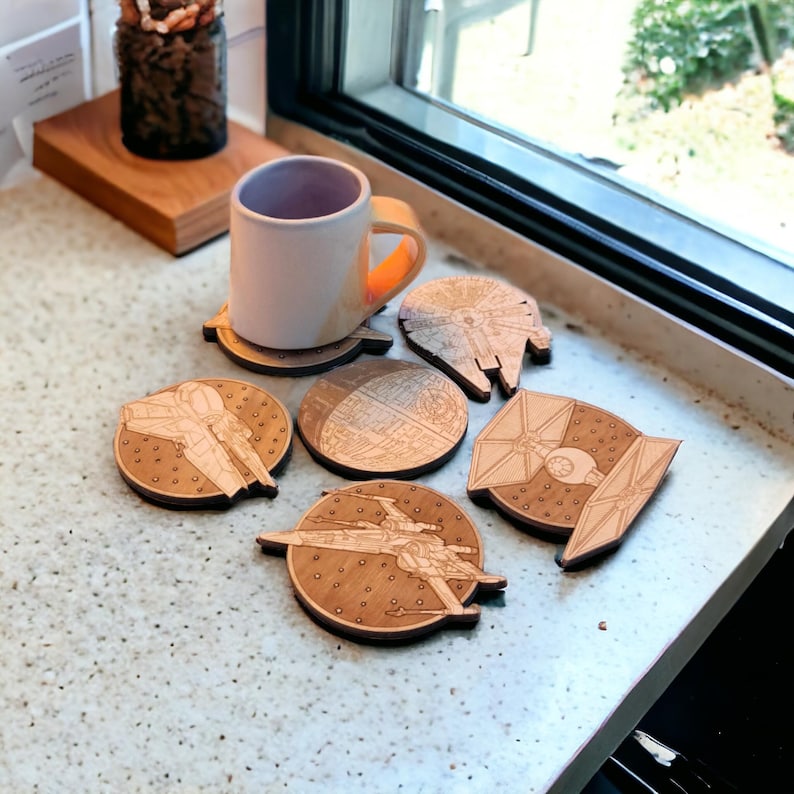 May include: A collection of wooden coasters featuring designs from a popular science fiction franchise. The coasters depict various spacecraft and space stations, with a mug resting on one. The coasters are made of light-colored wood.