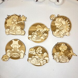 Pode incluir: Seis recortes de madeira circulares com personagens de videojogos em karts. Os desenhos cortados a laser são detalhados e a madeira tem uma cor castanha clara natural. Os personagens incluem uma princesa, Mario, Bowser e Toad.