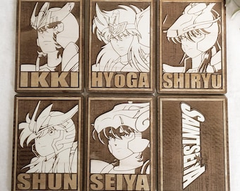 May include: Six wooden plaques with engraved images of characters from the anime series Saint Seiya. Each plaque features a different character, including Ikki, Hyoga, Shiryu, Shun, Seiya, and the title "Saints Seiya".