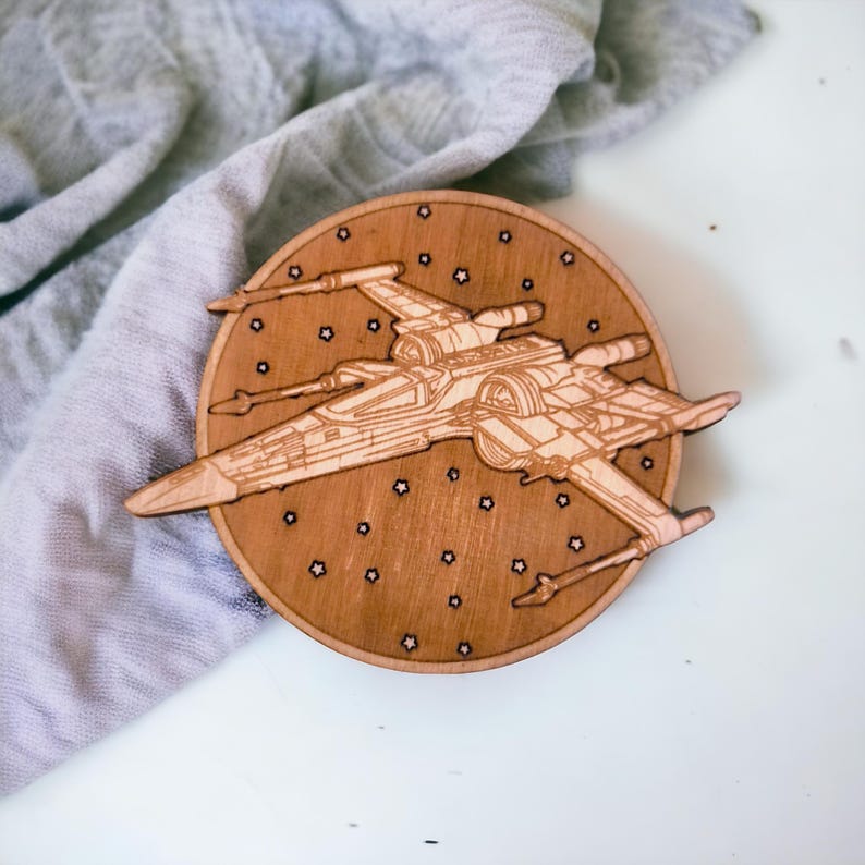 May include: A wooden coaster featuring a detailed laser-cut design of an X-wing starfighter. The coaster is circular, with a brown finish and small star-shaped cutouts around the starfighter. A great gift for fans of space movies.