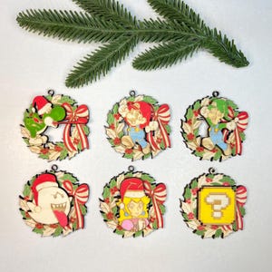 May include: A collection of six wooden Christmas decorations featuring characters from a popular video game series. Each ornament is a circular wreath with a red bow and holly. Characters include Yoshi, Mario, Luigi, Boo, Princess Peach, and a question block.