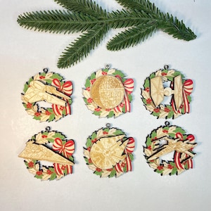 Op de afbeelding: Zes houten kerstversieringen met Star Wars-ontwerpen. Elke ornament heeft een lasergesneden ruimteschip of Death Star, geplaatst in een krans van hulst en rood lint. Een takje groenblijvende plant staat op de achtergrond.